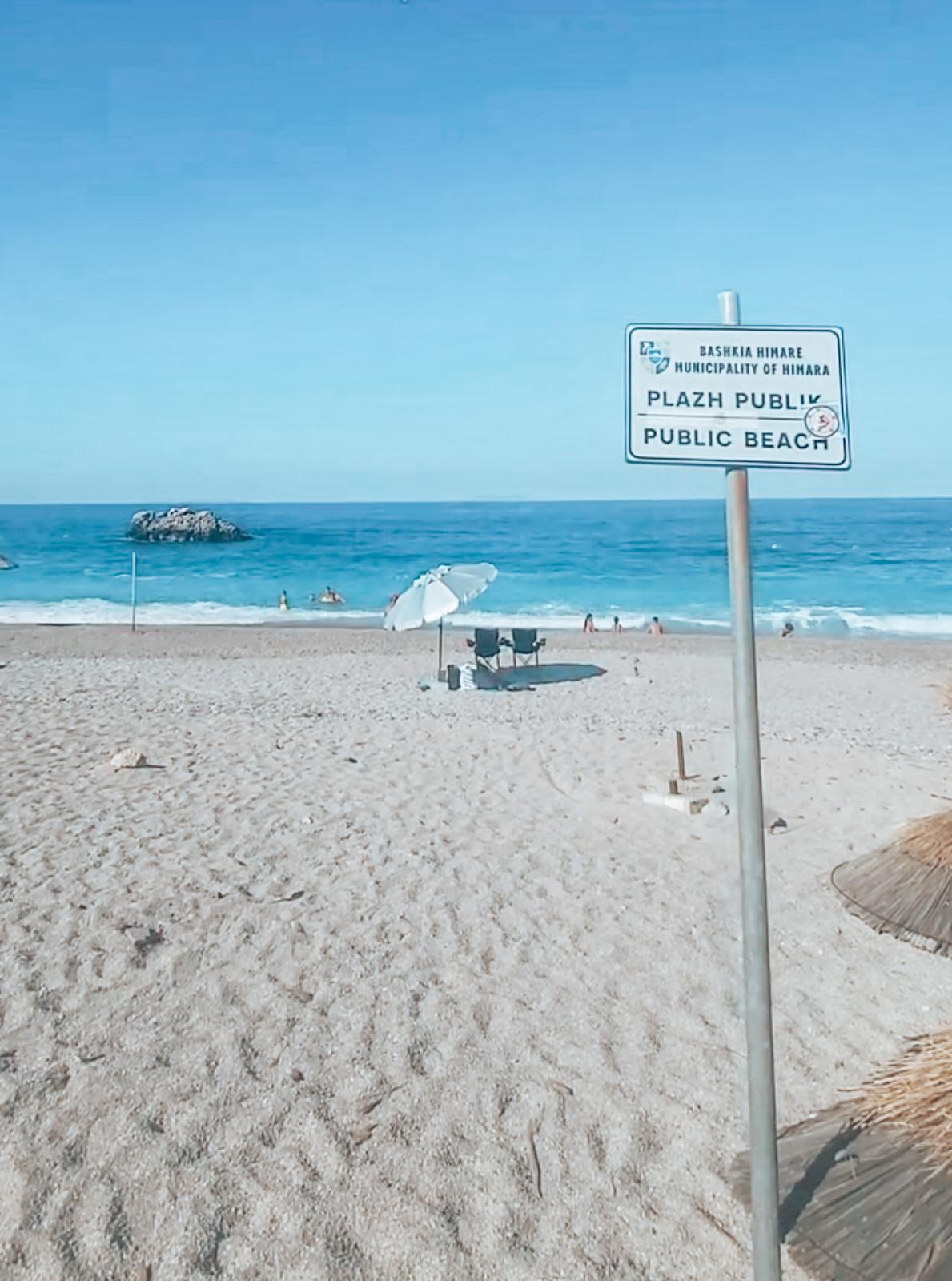 Livadhi Public Beach with rows of umbrellas, swimmers enjoying the calm water, and the open bay stretching toward the surrounding hills.