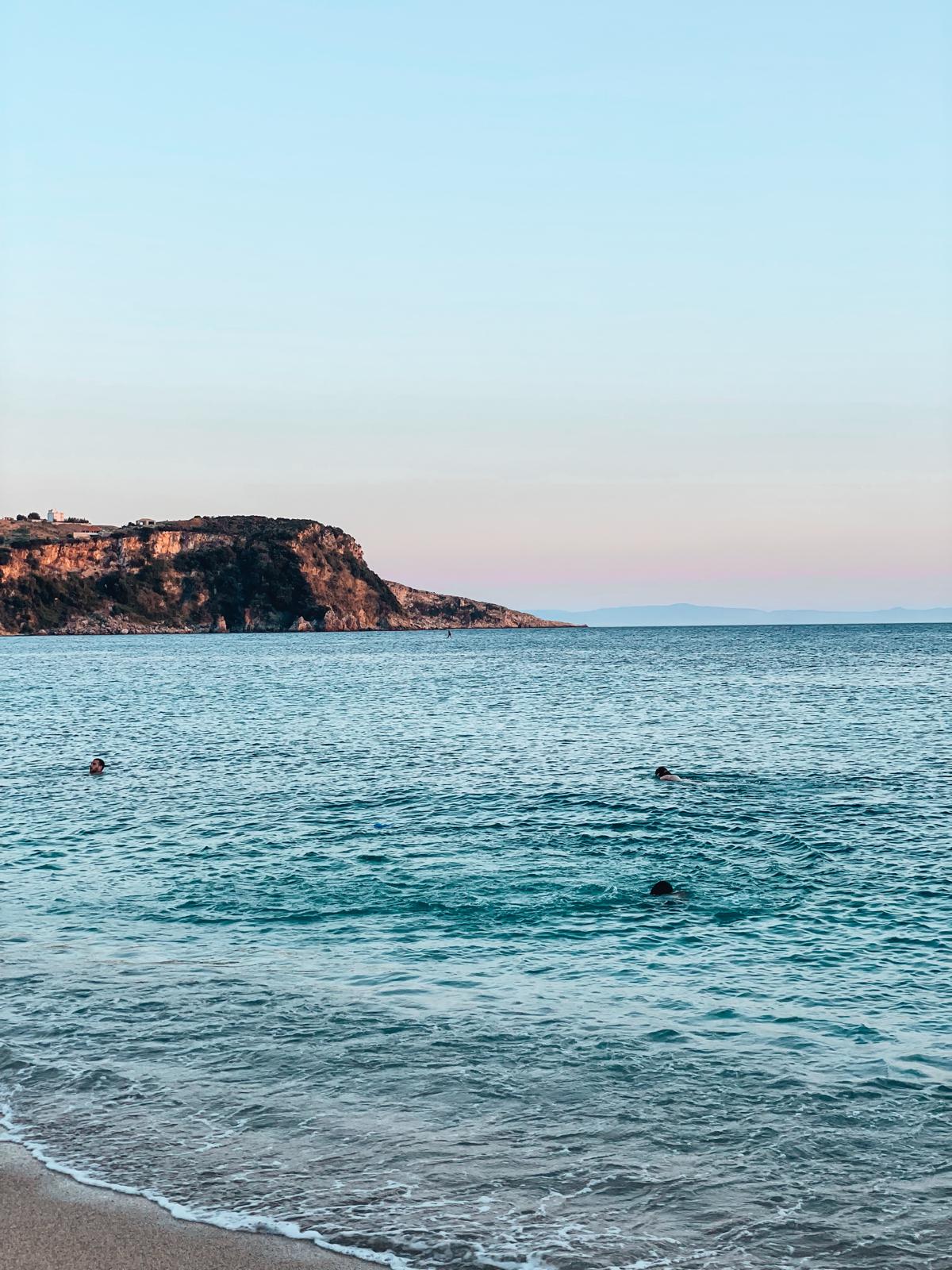 Clear, calm blue-green water washes gently over a pebbled shoreline at Potami Beach under bright sunlight. The simple, open seascape highlights the natural beauty of the Himara coastline near popular spots where visitors often look for parking in Himare.