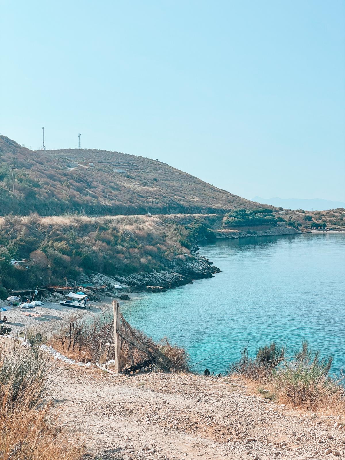 View from a parking area overlooking Potami Beach, where the shoreline and clear blue water are visible just beyond a small roadside stopping point. The scene shows the close proximity between parking spaces and the beach, with the sea stretching out under bright coastal light.