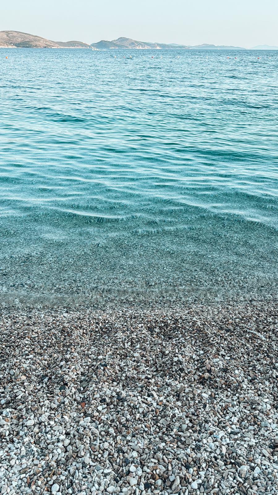 Sunny day at Saranda Beach with calm water and a promenade lined with buildings in the background. The scene highlights one of the area's popular spots for swimming and seaside relaxation.