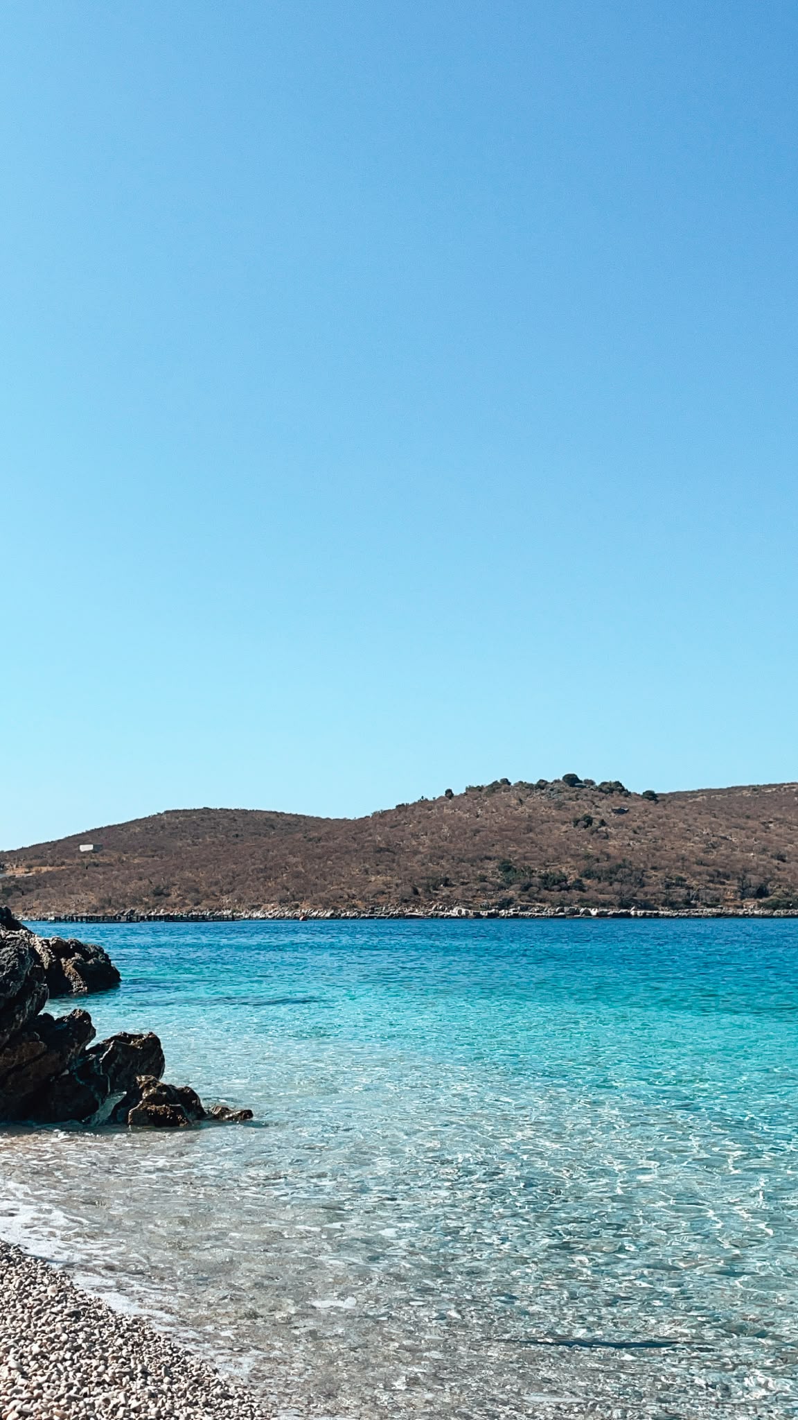 Potami Beach with a wide stretch of pebbled shoreline, clear blue-green water, and gentle waves meeting the coast under a bright sky. The peaceful natural setting reflects why it is considered part of the Best Beaches in Himare.
