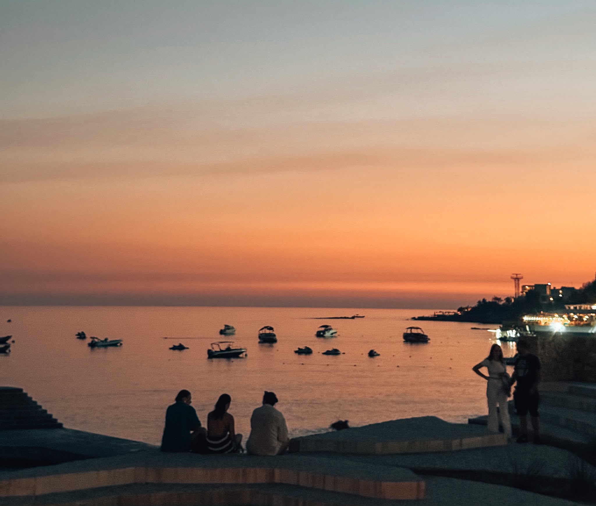 Dhermiu Beach at sunset with warm orange and pink tones spreading across the sky and reflecting on the calm Ionian Sea. The shoreline is dotted with soft waves and silhouettes of beach terraces and loungers as the light fades over the coast. The scene captures a peaceful evening atmosphere that fits the Best Beaches in Himare.