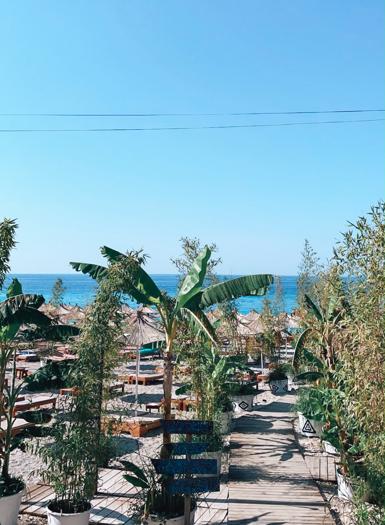 Livadhi Beach in Himarë with a long stretch of shoreline bordered by clear blue water and green hills in the background. The beach is one of the beaches in Himarë reachable by scooter and offers expansive coastal views along the Albanian Riviera.