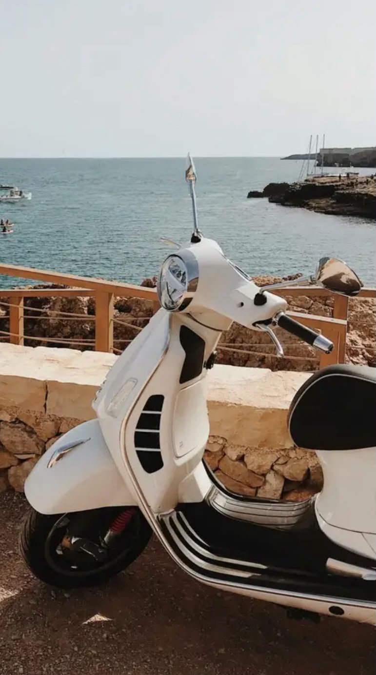 A scooter parked or riding along the Himarë lungomare promenade beside the sea, with the coastal road running parallel to the waterfront and surrounding hills in the background. The scene reflects easy access between town and nearby beaches in Himarë reachable by scooter along the Albanian Riviera.