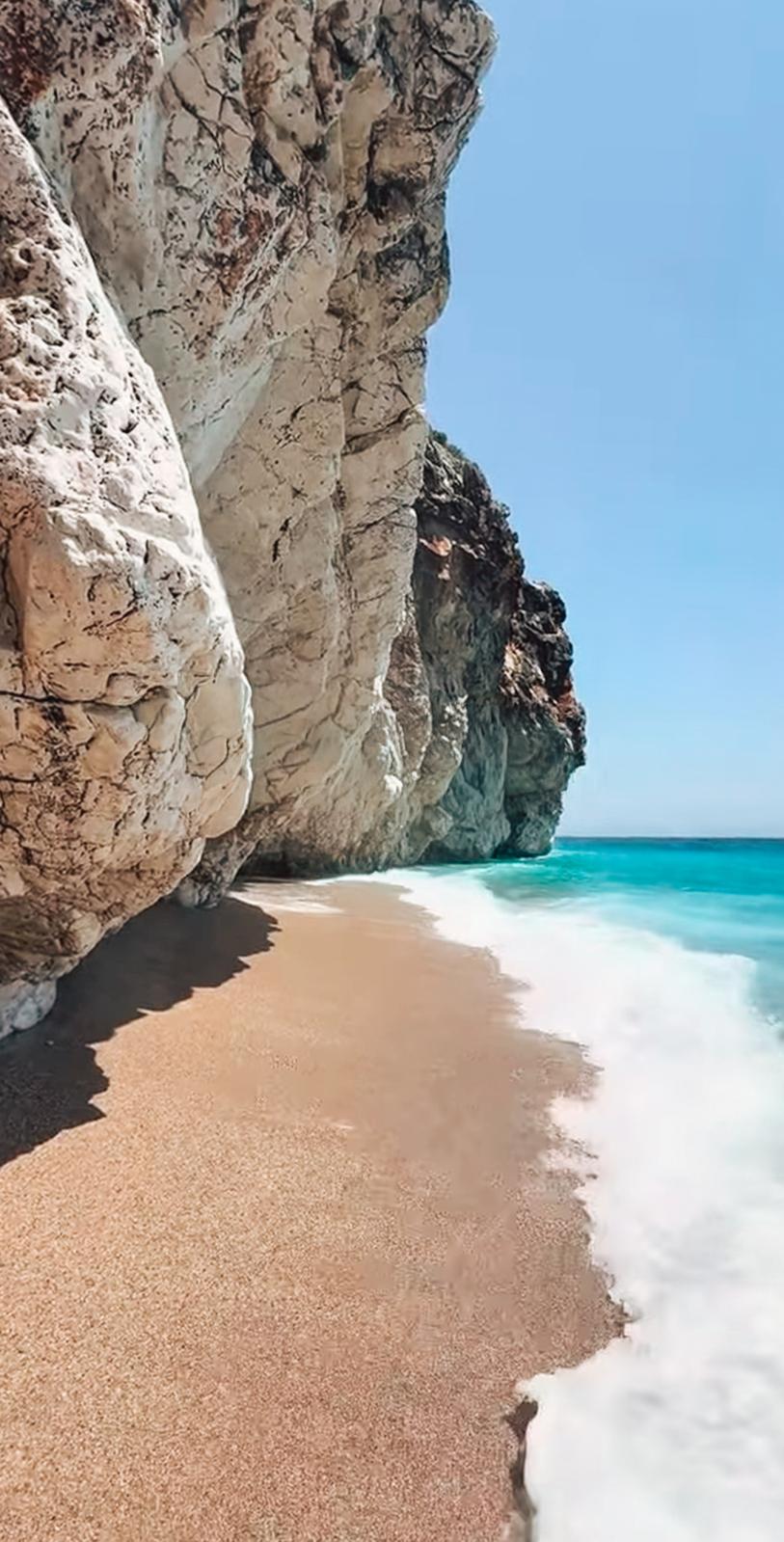 Gjipe Beach with clear turquoise water meeting a pebble shoreline at the mouth of Gjipe Canyon. Visitors relax along the coast surrounded by steep cliffs, highlighting the destination reached when following the route from Himarë to Gjipe Beach.