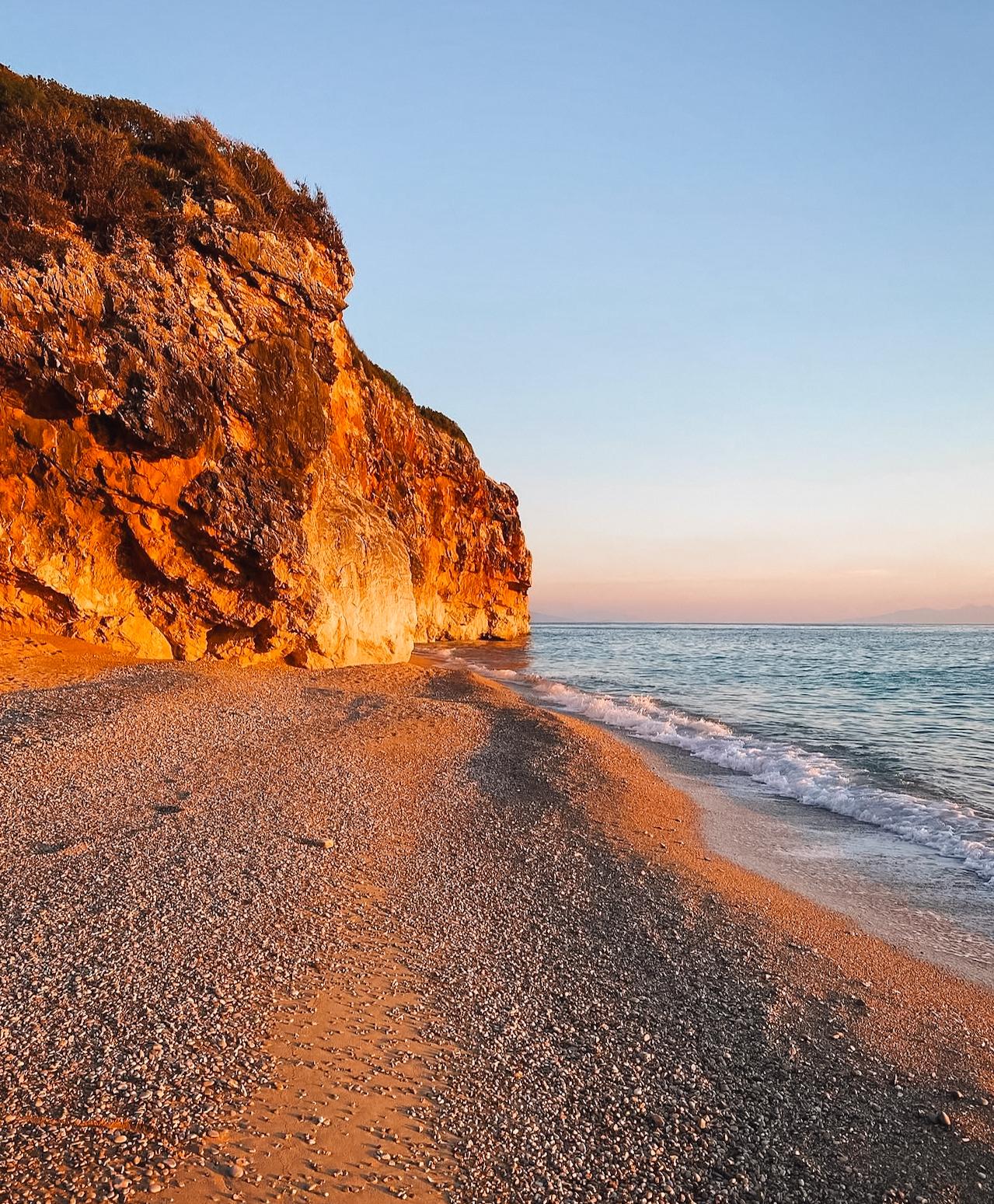 Sunset over Gjipe Beach as golden light reflects across the calm sea and illuminates the surrounding cliffs. The coastal landscape creates a peaceful evening scene at one of the most popular stops for travelers visiting Gjipe Beach from Himarë.