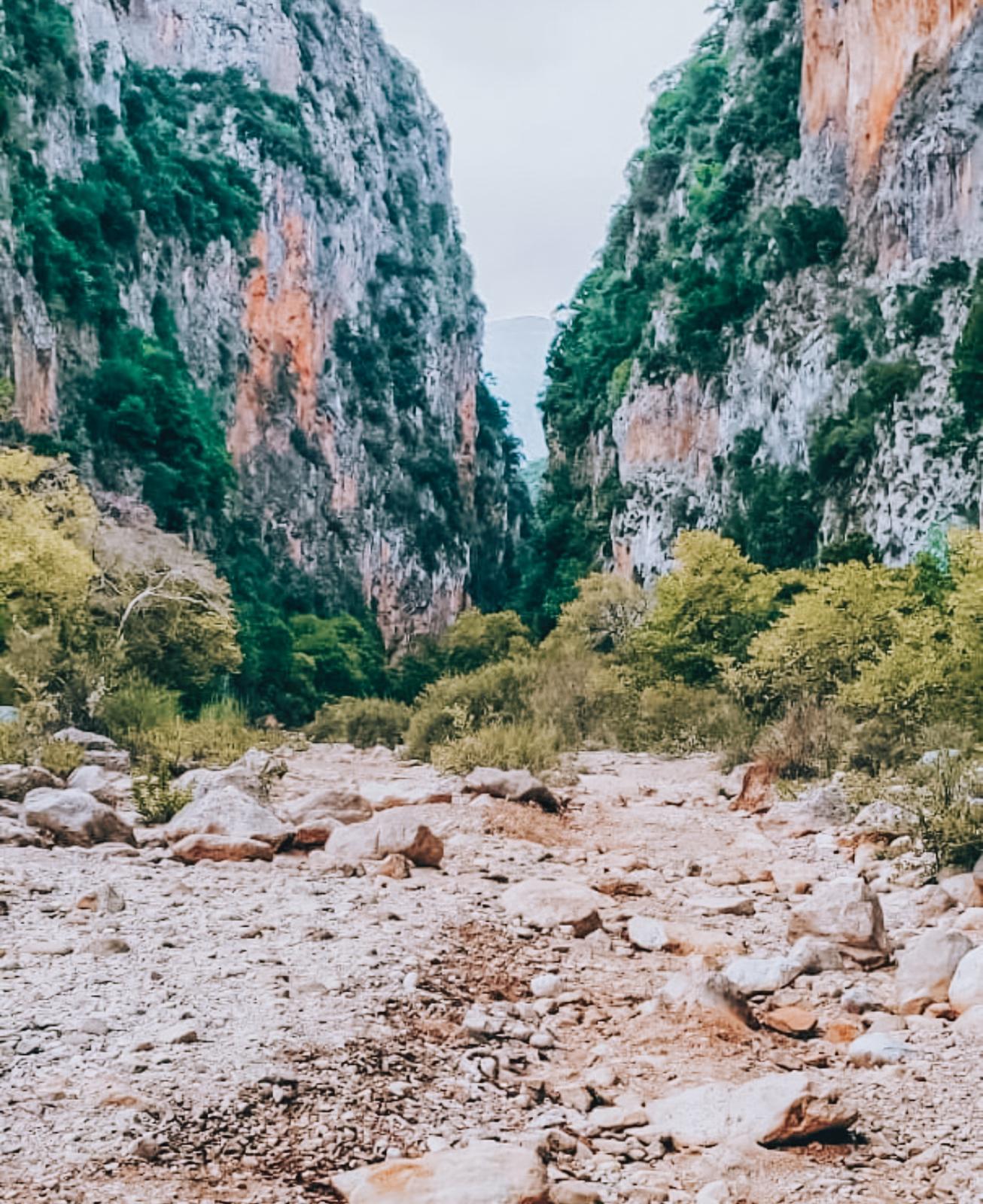View of Gjipe Canyon from the sandy and pebbled shoreline of Gjipe Beach, where steep limestone cliffs frame the narrow gorge opening toward the sea. The scene shows the dramatic landscape that surrounds visitors after completing the walk from Himarë to Gjipe Beach.