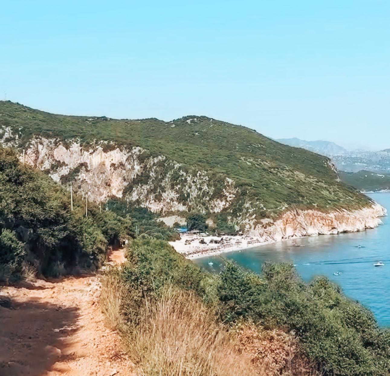 View of Gjipe Beach from the walking road between the parking place and the shoreline. The turquoise water curves between steep rocky cliffs, showing the final approach visitors take when following directions on how to get to Gjipe Beach from Himarë.
