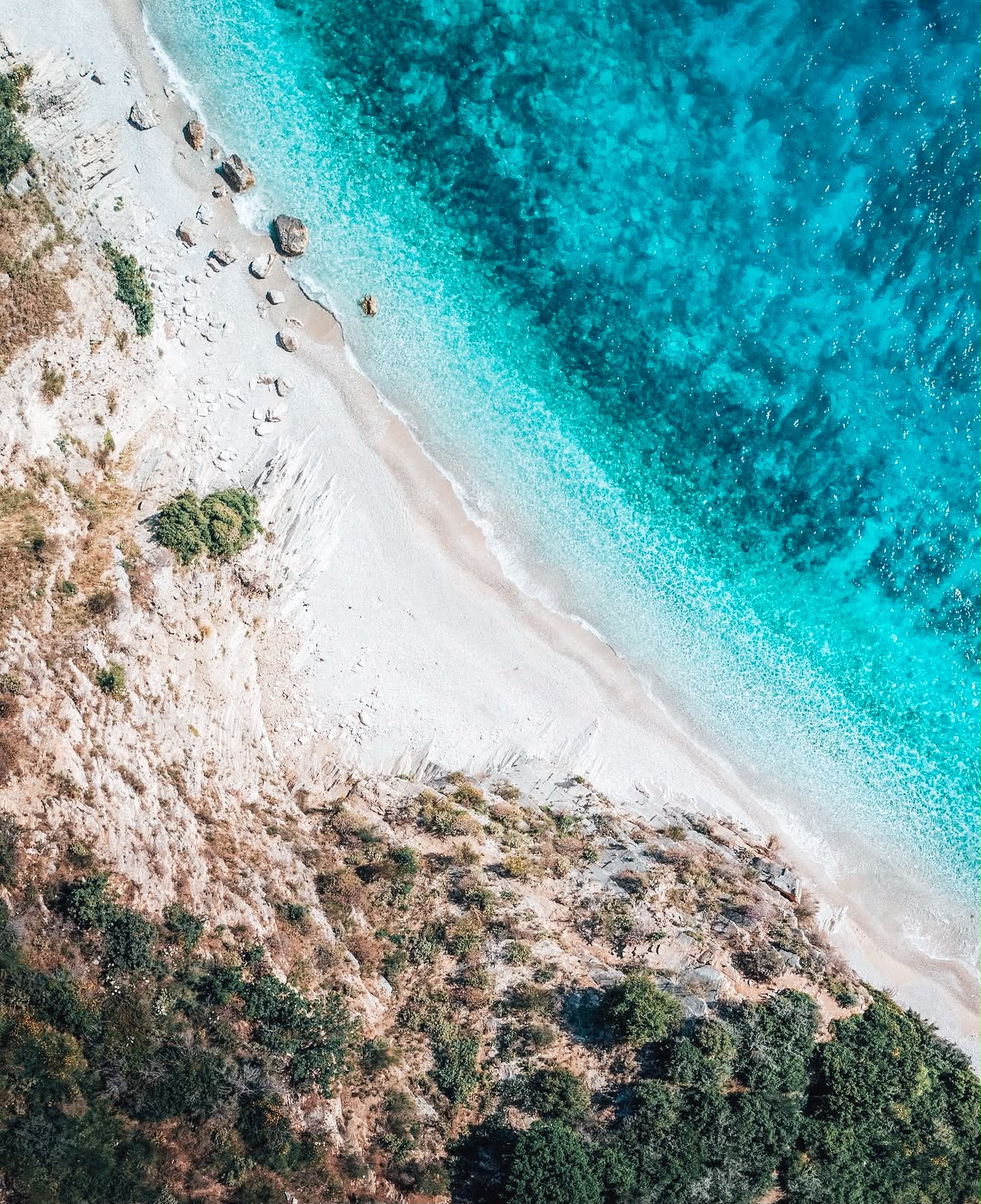 Aerial drone view of Filikuri Beach showing a hidden crescent of shoreline tucked between steep cliffs and deep blue sea. The secluded layout emphasizes the untouched nature of the coast, aligning with the idea of the best quiet beaches near Himarë you can walk to.