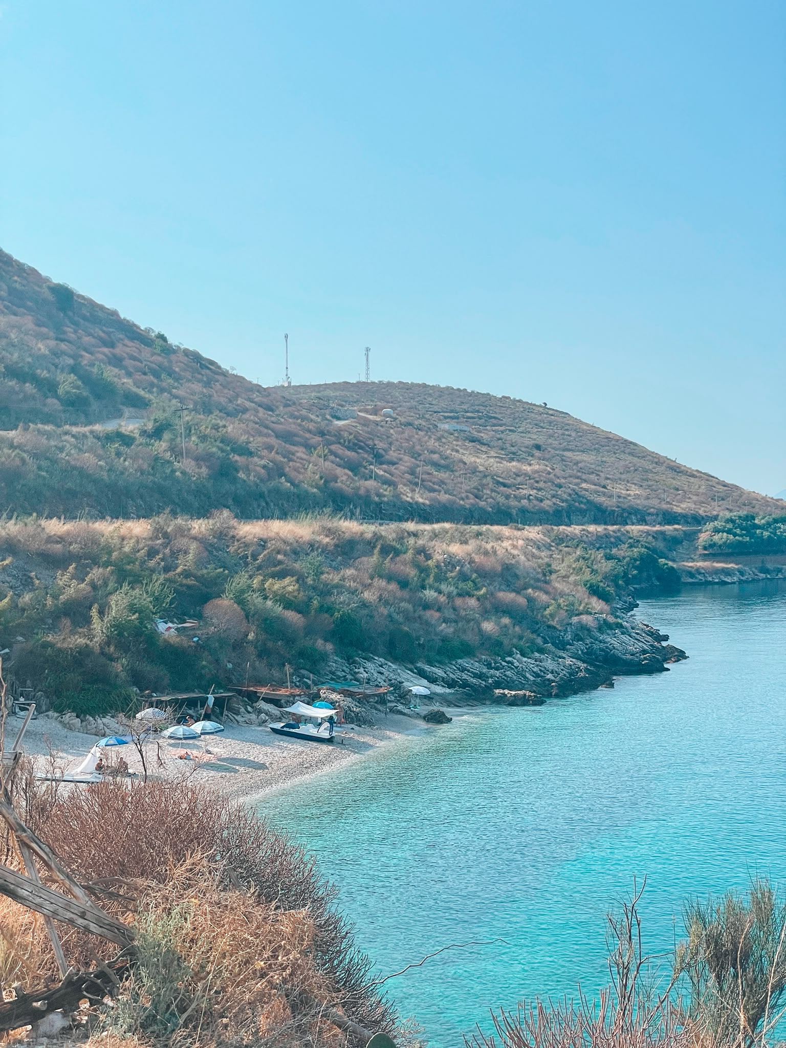 Potami Beach with a long stretch of pebbled shoreline meeting clear blue water, framed by rocky cliffs and scattered greenery. The calm coastal setting gives a sense of a more relaxed escape compared to busier nearby beaches during peak season.