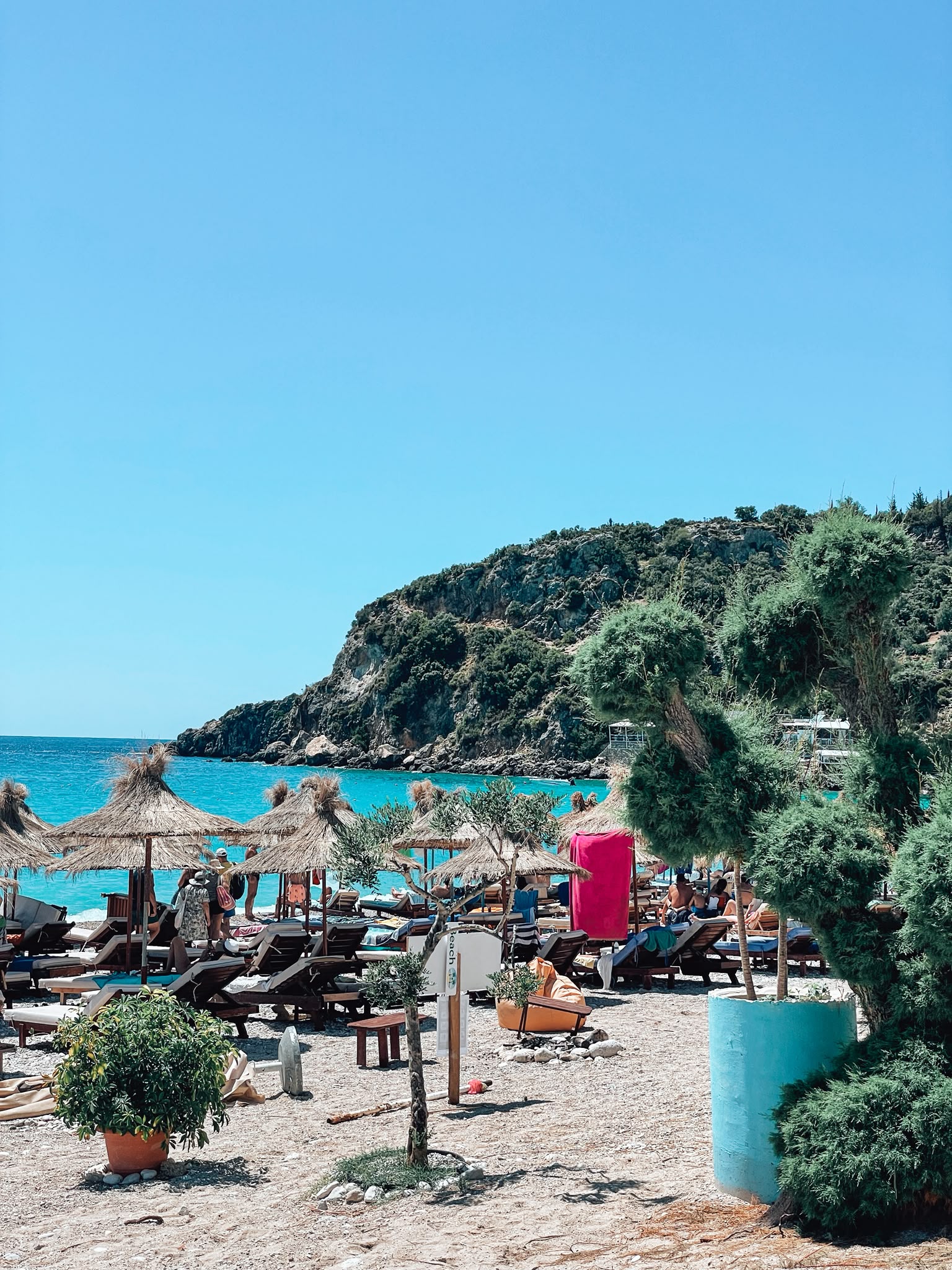 Jala Beach with clear turquoise water, a wide stretch of pebbles, and sun loungers along the shore as visitors enjoy a sunny summer day. The lively beach scene highlights one of the most popular coastal spots near Himara during peak season.