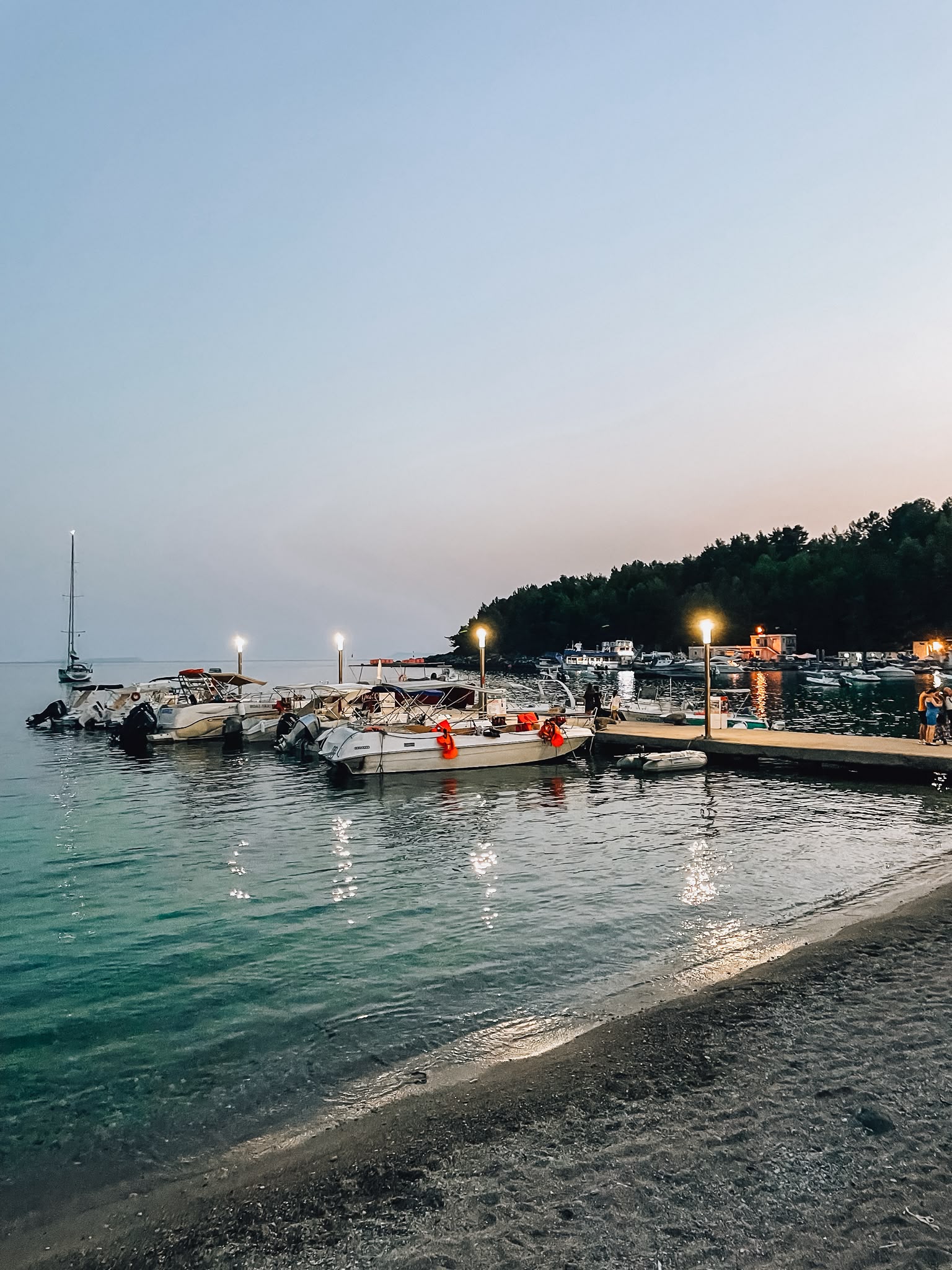 Spile Beach at sunset with warm golden light reflecting across the calm sea as people stroll along the shoreline. The peaceful evening atmosphere shows why Himara is worth visiting in peak season while highlighting one of its most popular beaches.