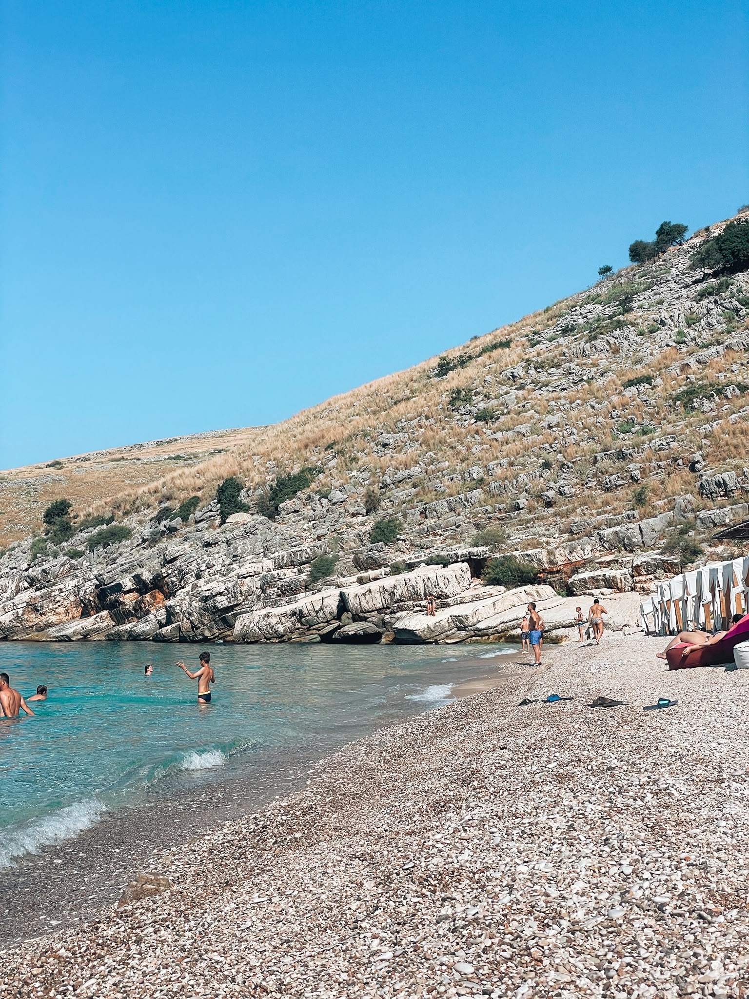 Llamani Beach with a curved bay of turquoise water meeting smooth pebbles and steep green hills rising behind it. The sheltered setting creates a quiet, scenic atmosphere that stands out even during the busy peak season near Himara.