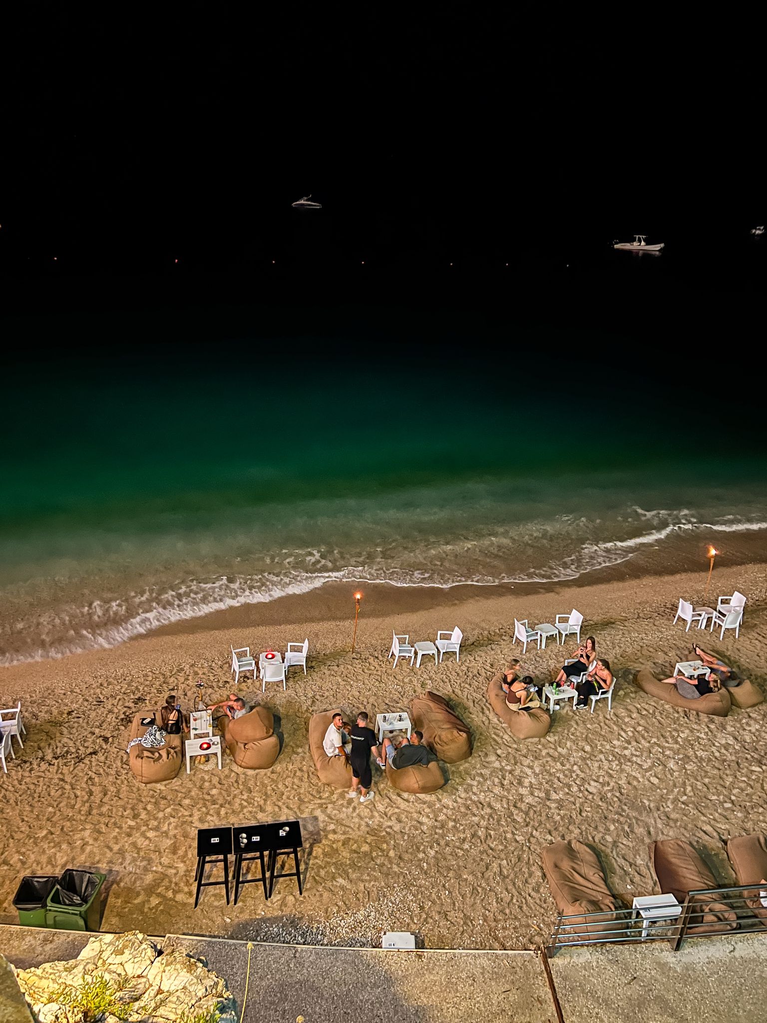 Himara beach at night with dark, gently rippling water reflecting nearby lights from the shoreline and promenade. The calm evening atmosphere highlights the peaceful seaside setting that makes staying near the beach in Himara especially appealing.