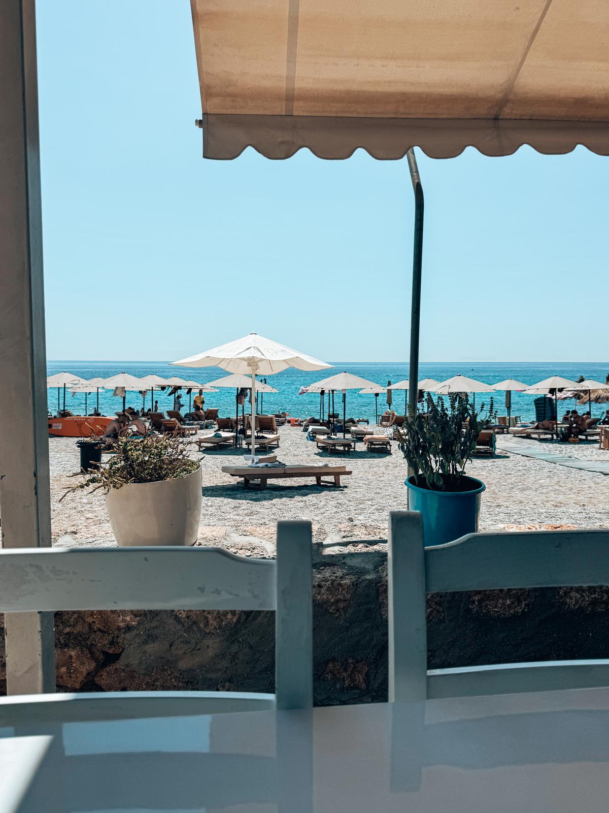 View of Livadhi Beach from the beachfront restaurants overlooking the turquoise sea, sun loungers, and the pebbled shoreline below.