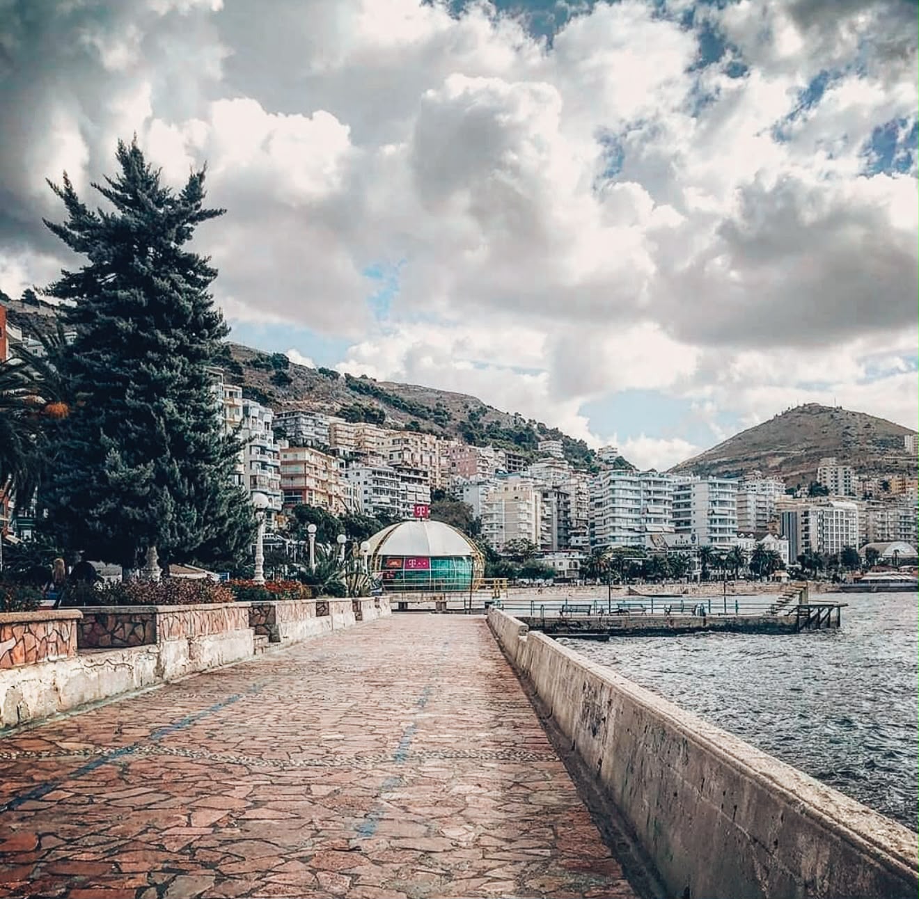 Palm-lined promenade along the Sarandë waterfront with the Ionian Sea stretching beside it. People stroll along the seaside walkway as cafés and buildings line the shore, capturing the lively atmosphere of the city.
