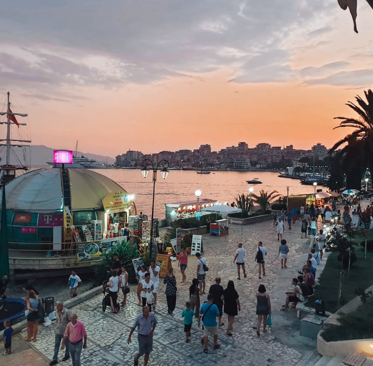 View along the Sarandë promenade overlooking the city, with palm trees lining the waterfront and buildings climbing the hillside above the Ionian Sea. The seaside walkway is busy with people enjoying the waterfront atmosphere.