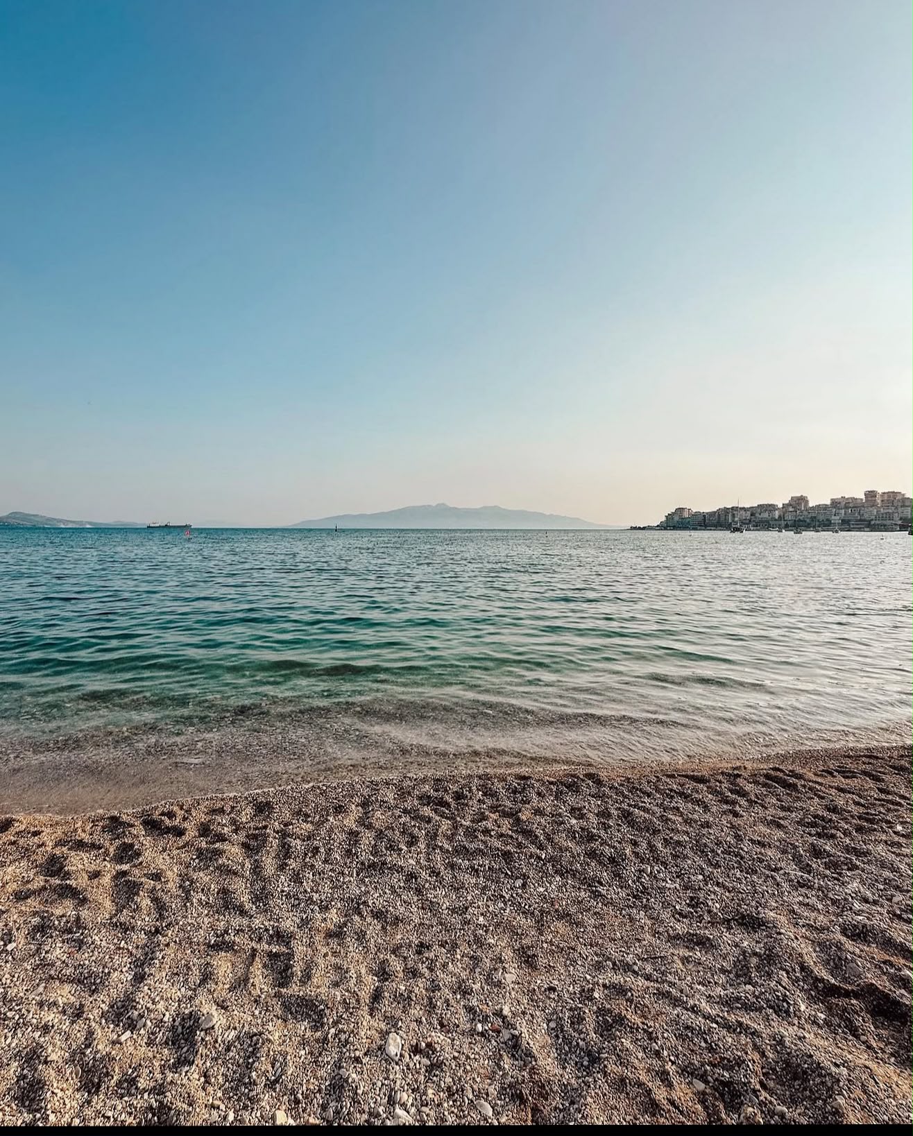 Sandy beach in Sarandë with clear turquoise water gently meeting the shore along the Ionian Sea. Sun loungers and umbrellas line the beach as visitors relax against a backdrop of the coastal town and nearby hills.