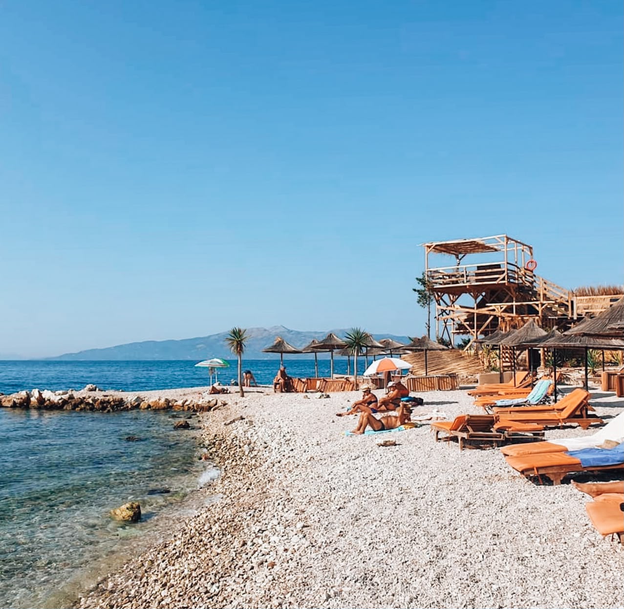 peblee beach near the Port of Sarandë with clear turquoise water and rows of sun loungers facing the Ionian Sea. Boats are visible nearby as people relax and swim close to the city waterfront.