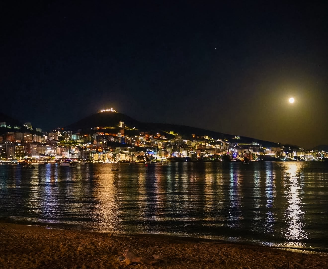 Night view of Sarandë with the promenade softly lit by streetlights and reflections shimmering across the calm Ionian Sea. City lights climb the hillside, creating a warm glow along the waterfront.