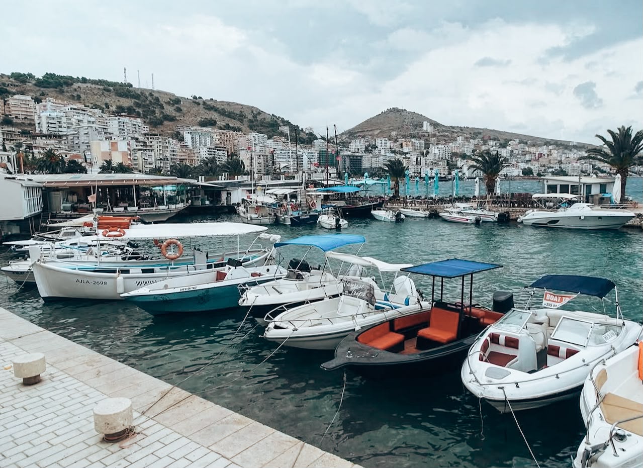 View of the Port of Sarandë with ferries and boats docked along the waterfront, backed by the city's hillside buildings. The busy harbor highlights Sarandë as a gateway to the Albanian Riviera and nearby islands.