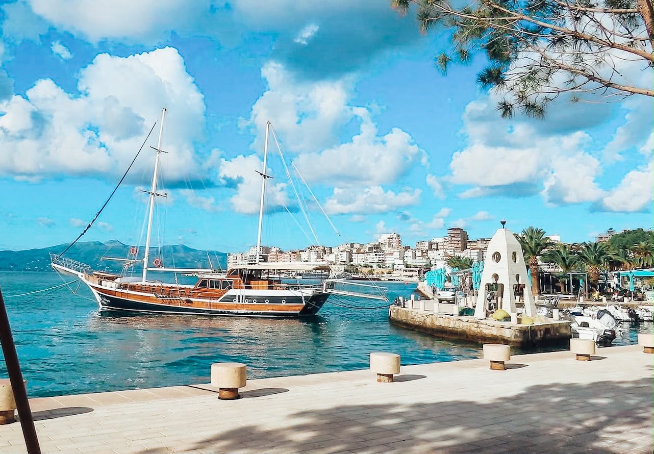 Wide view from the Sarandë promenade looking over the harbor and turquoise Ionian Sea. The waterfront stretches along the city with palm trees, boats, and hillside buildings creating a layered coastal cityscape.