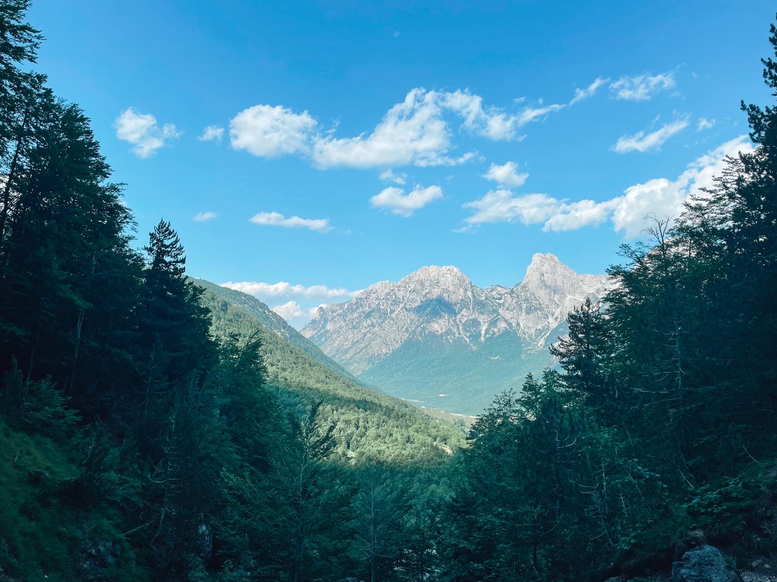Rugged peaks of the Albanian Alps rise above a green mountain valley with dramatic rocky slopes and clear skies. The landscape highlights the breathtaking scenery travelers can experience when visiting Valbona and Theth beyond the hiking trails.