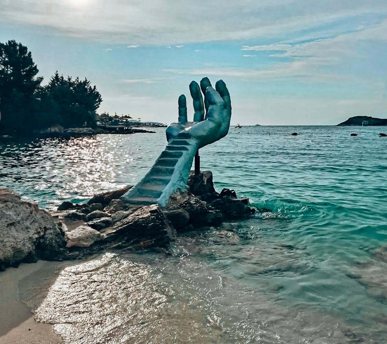 Giant wooden hand sculpture in Ksamil overlooking turquoise water, with visitors posing for photos against the scenic coastal backdrop. This popular photo spot is often visited alongside the beaches when exploring Are There Free Beaches in Ksamil.
