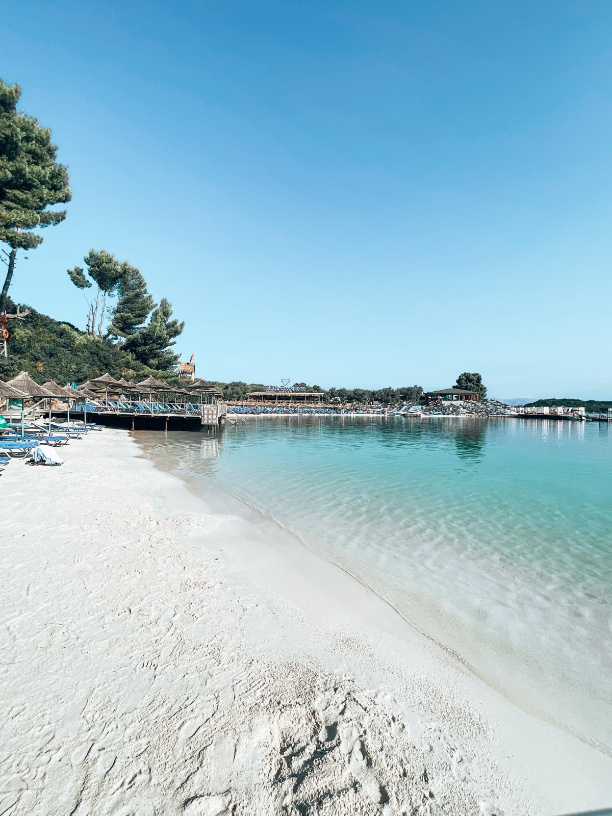 White sand beach in Ksamil with shallow turquoise water, sunbathers relaxing along the shore, and small islands visible in the distance. This scenic coastline is a great example of the beaches visitors discover when researching Are There Free Beaches in Ksamil.