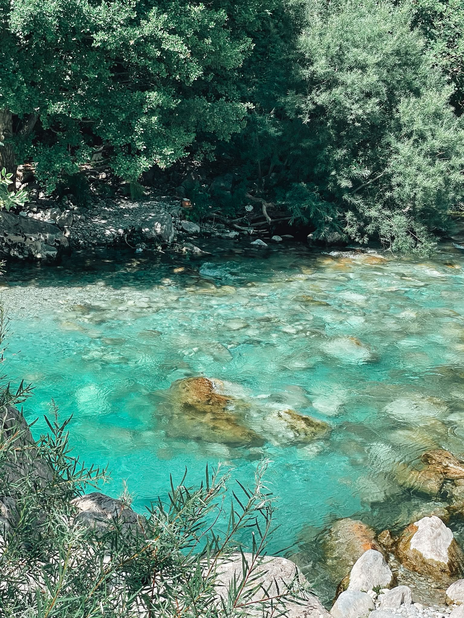 Crystal clear turquoise water flows through the Valbona River, with rocky banks and the rugged peaks of the Albanian Alps rising in the background. The river is one of the highlights of Valbona Valley and showcases the region's striking mountain scenery.