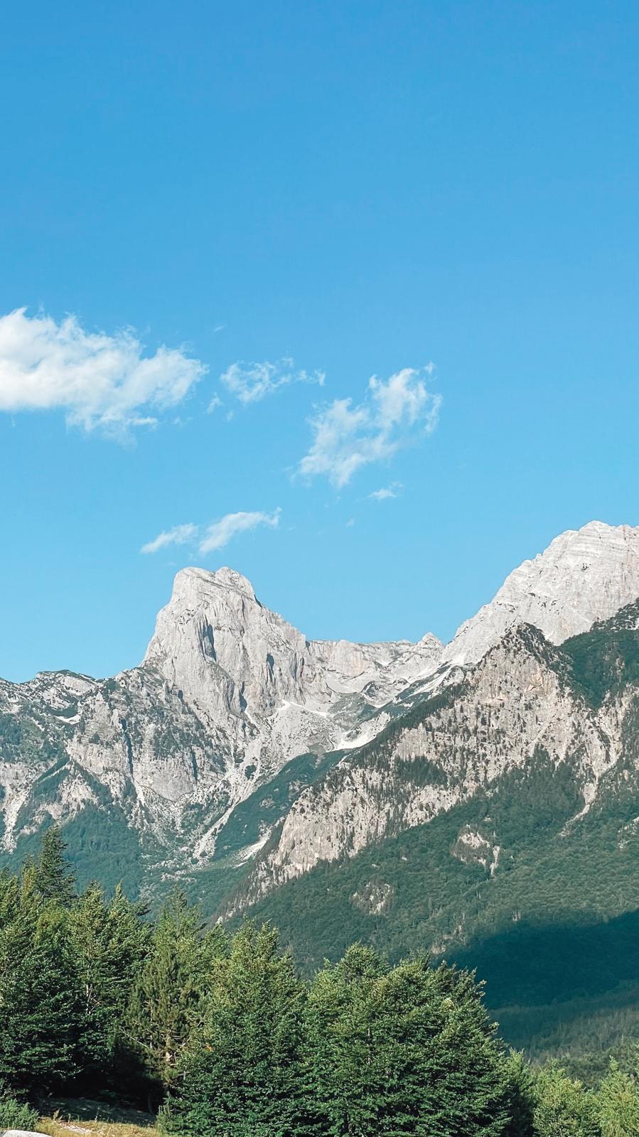 harp mountain peaks of the Albanian Alps stretch across the horizon above a peaceful valley with lush greenery and rugged terrain. The view captures the dramatic landscapes that make Valbona and Theth popular destinations for nature lovers.