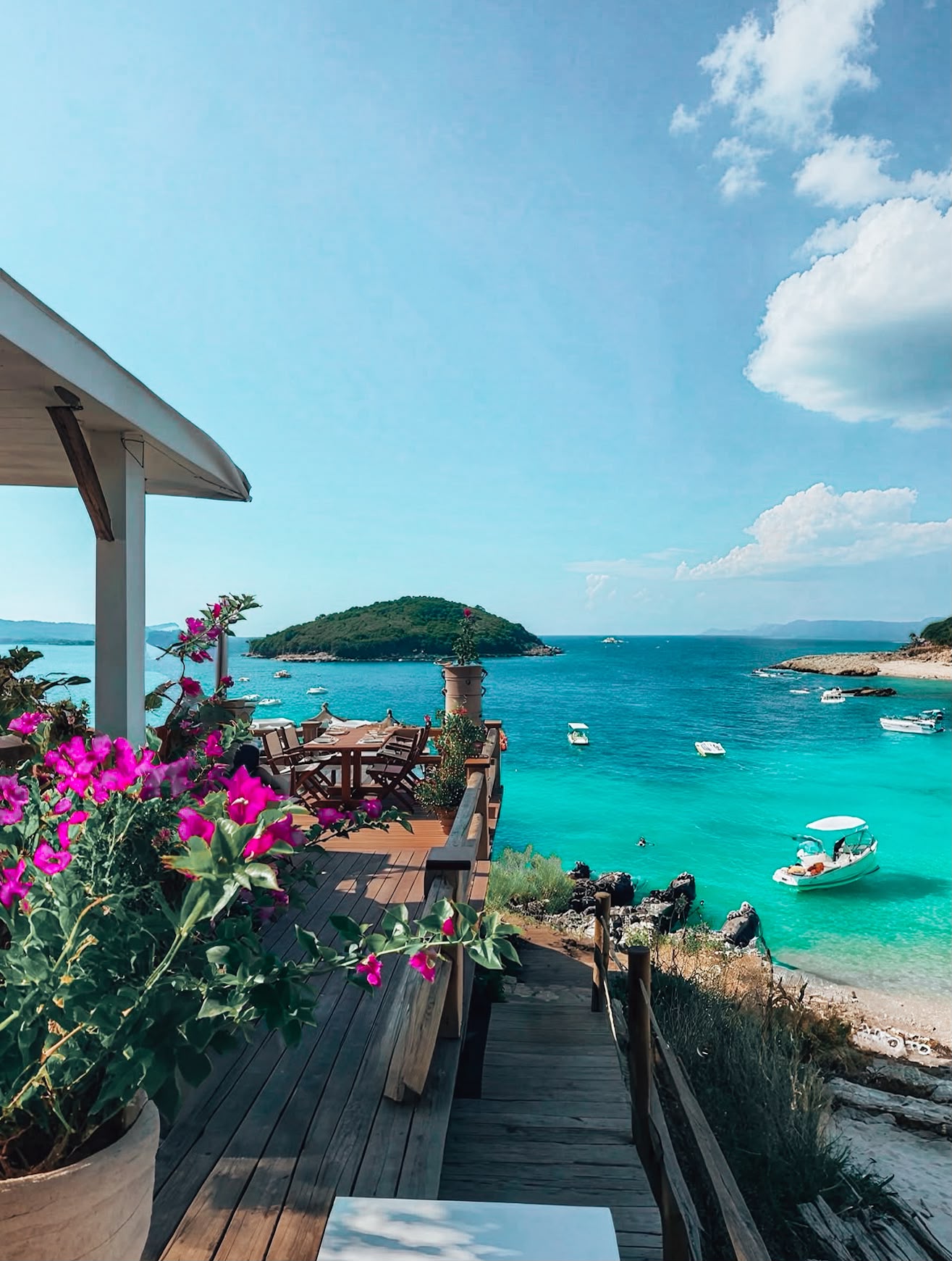 Coastal view of Ksamil from a restaurant terrace, showing turquoise sea water, nearby islands, and the shoreline stretching across the horizon. The relaxing dining viewpoint captures the scenery visitors can enjoy while exploring the area.