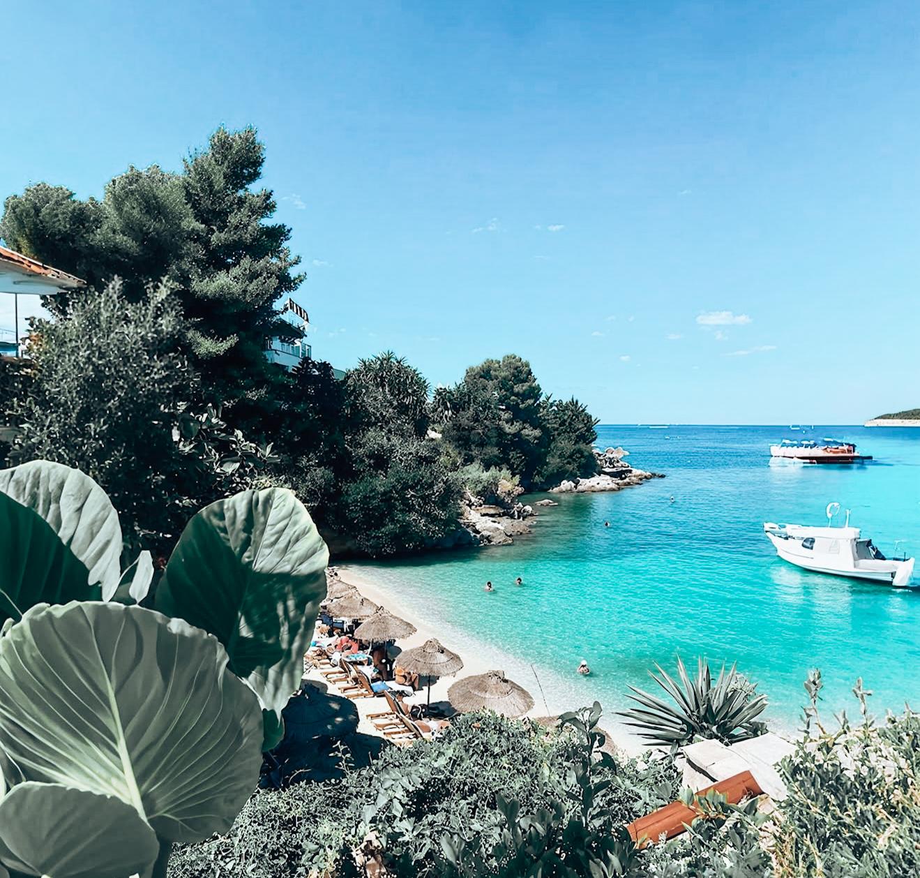 Ksamil beach viewed from a nearby street with turquoise water, sandy shoreline, and umbrellas along the coast. The street perspective shows the village setting and helps travelers compare Ksamil vs Saranda when choosing where to stay in Albania.