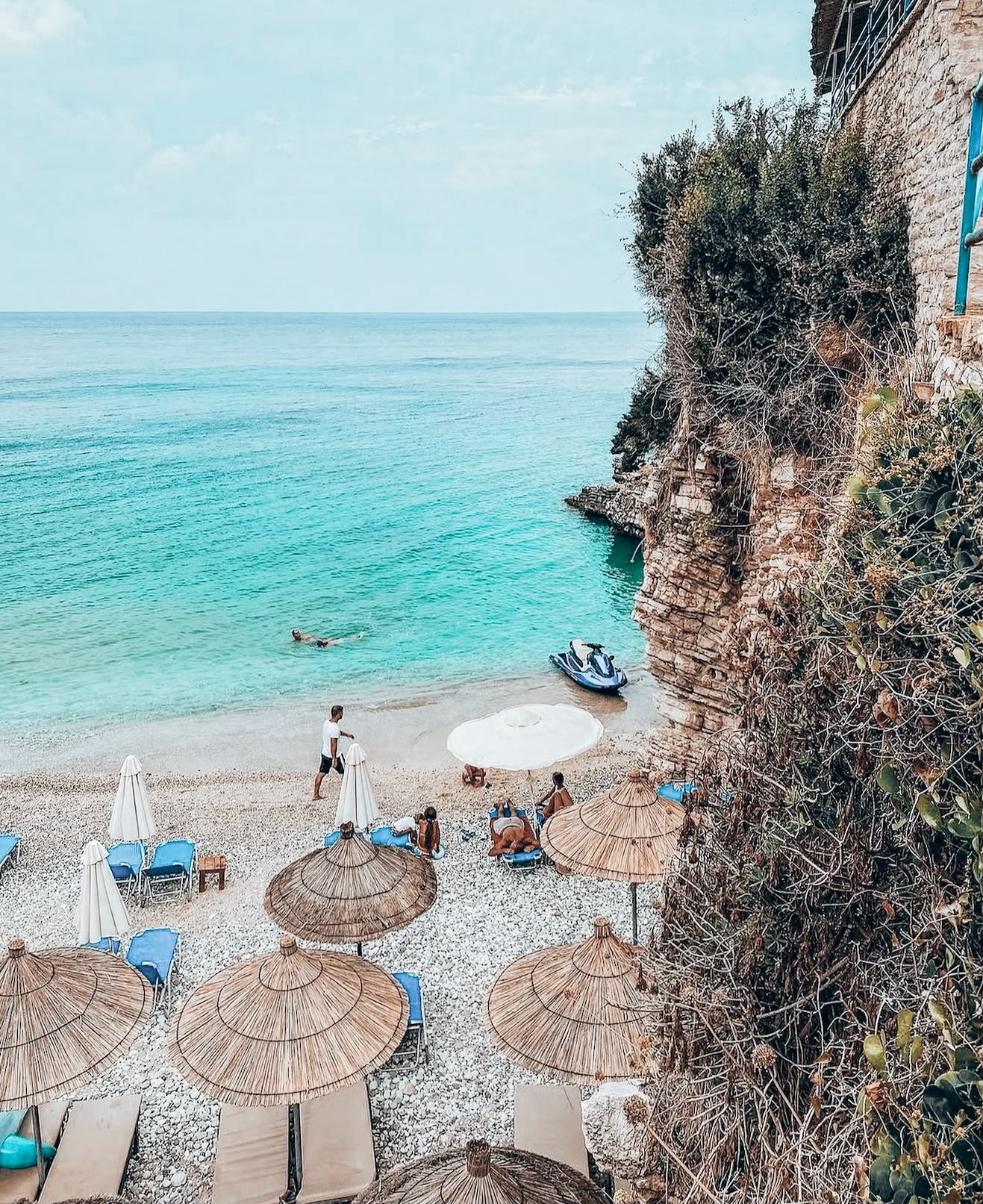 Clear turquoise water at Pulbardha Beach with rocky cliffs surrounding the shoreline and sun loungers set along the coast. The peaceful seaside landscape shows the natural beauty of the Ksamil area.
