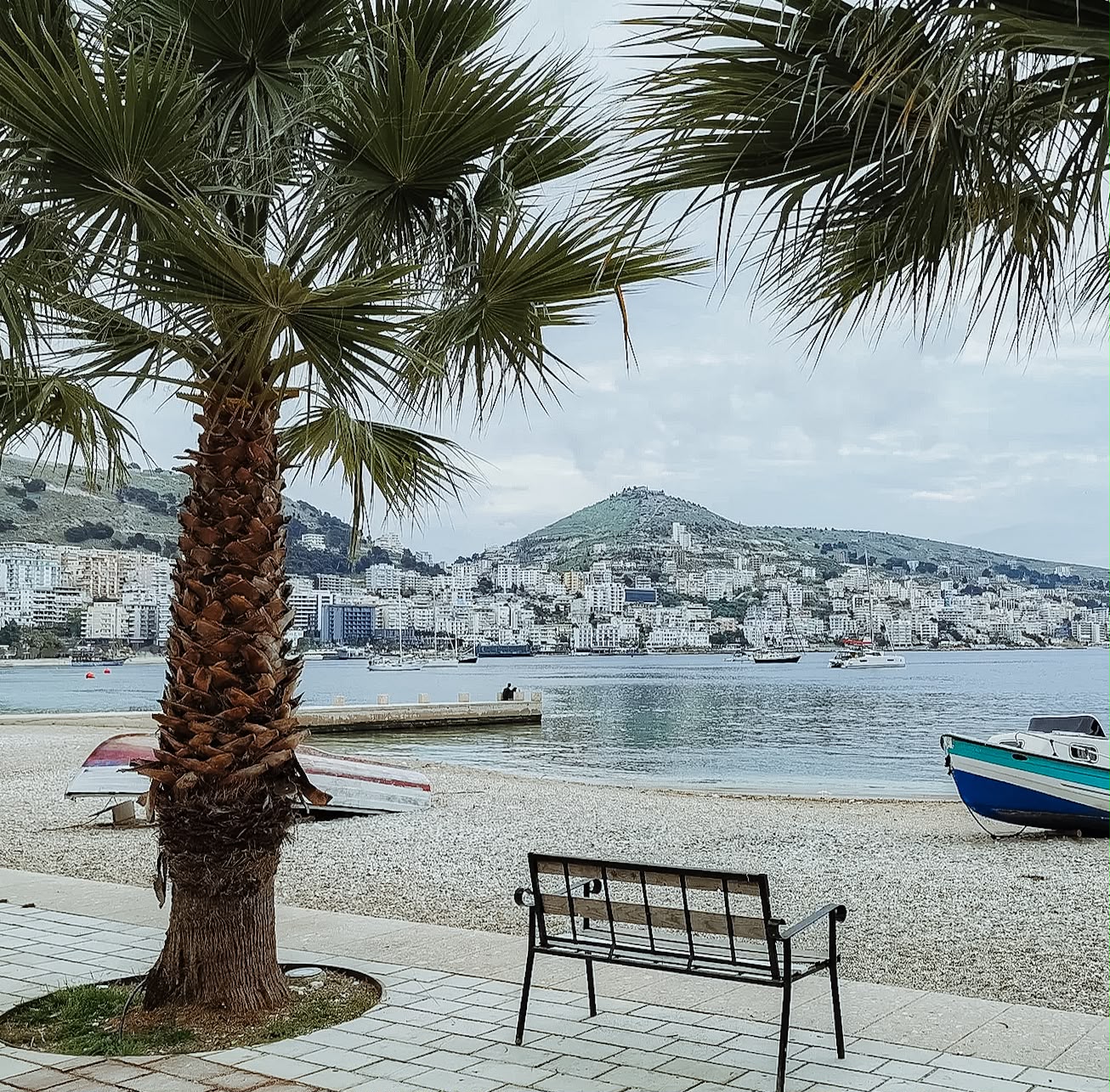 Saranda promenade along the waterfront with palm trees, seaside cafes, and views of the Ionian Sea. The coastal walkway shows the starting point for travelers planning how to get from Saranda to Ksamil.