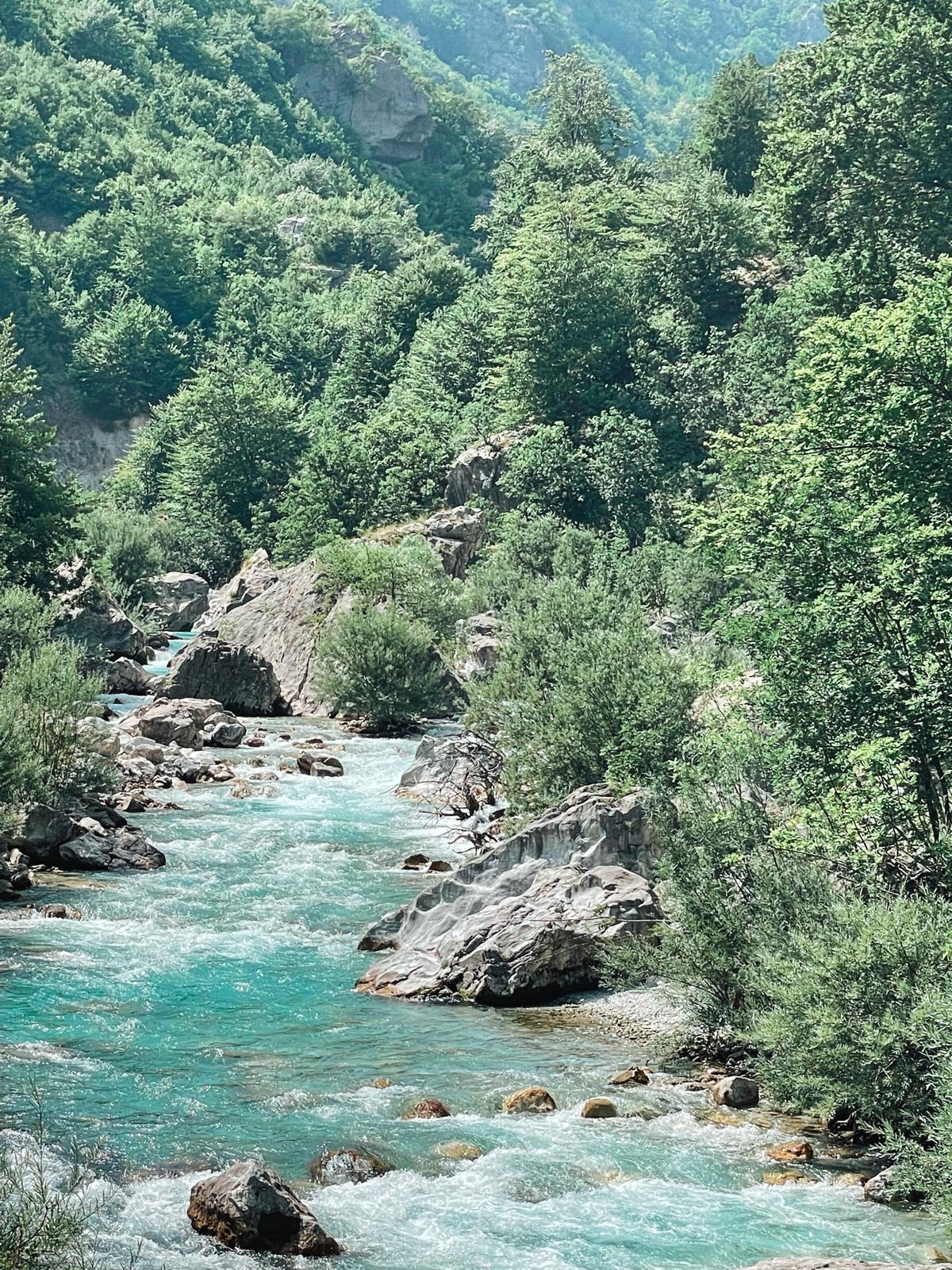 Clear turquoise water flows through the Valbona Valley with tall green mountains rising on both sides. The peaceful riverside landscape shows the natural beauty visitors can enjoy in Valbona even without hiking.